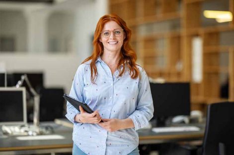 Woman with red hair and glasses standing in an open-plan office, holding a tablet