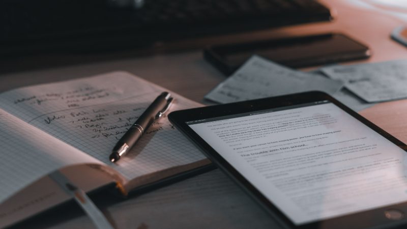 Handwritten notes in an open notebook with a pen, beside a tablet displaying an article